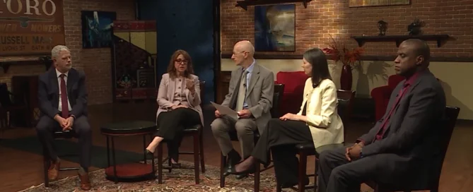 Five diverse professionals sitting in chairs during a broadcast panel discussion in a brick-walled studio.