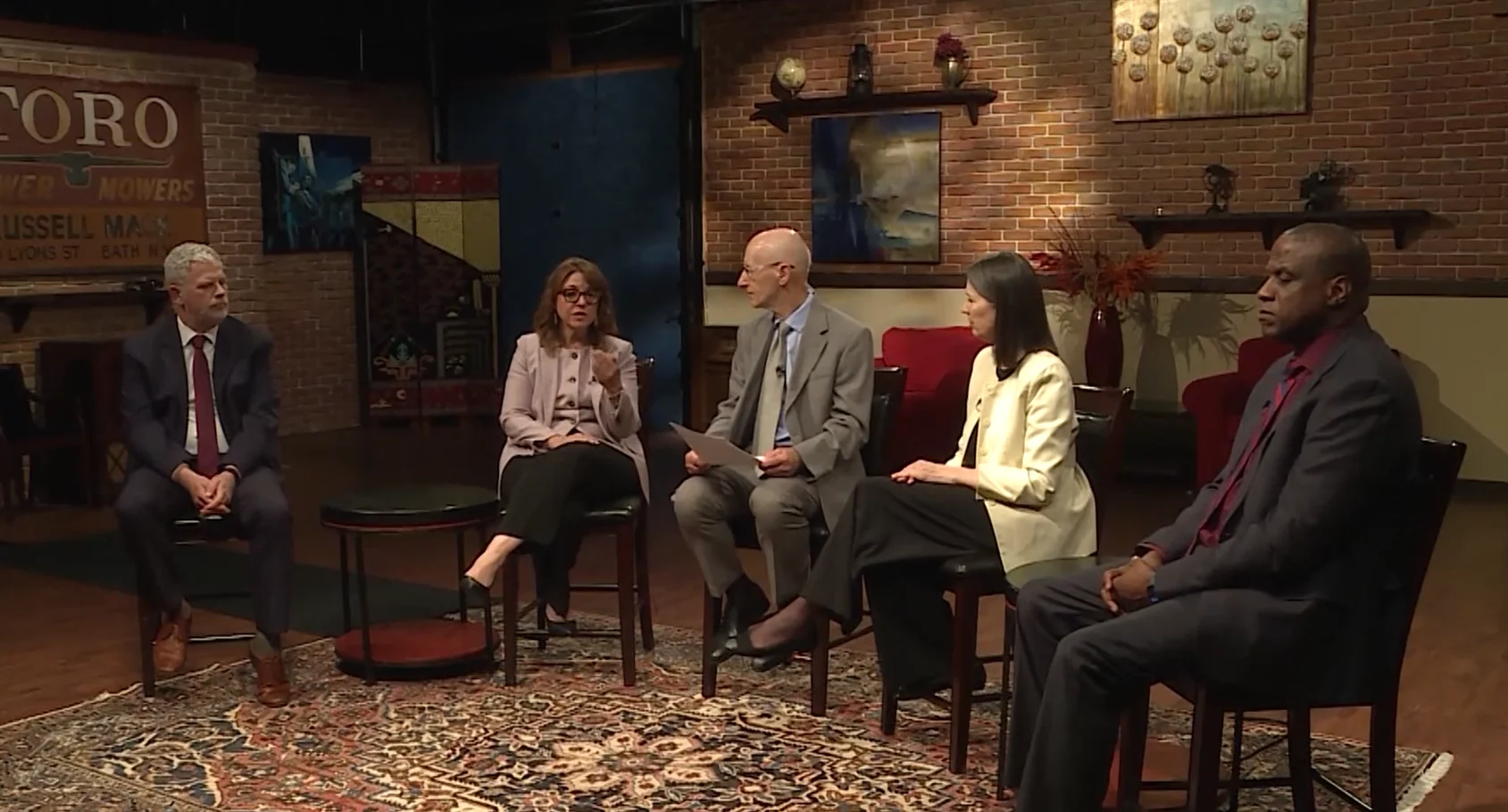Five diverse professionals sitting in chairs during a broadcast panel discussion in a brick-walled studio.